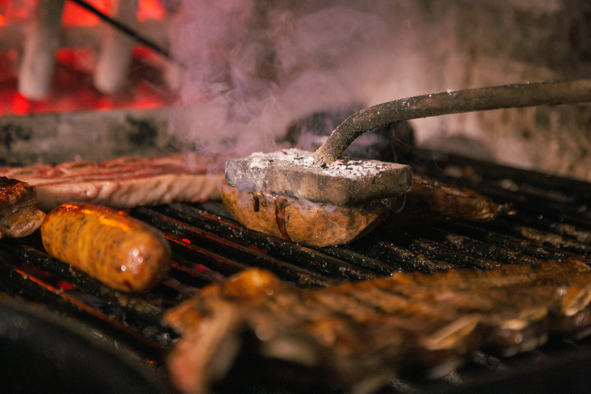 Una prensa de hierro apretando un corte sobre la parrilla, entre el humo, con chorizos y costillas alrededor