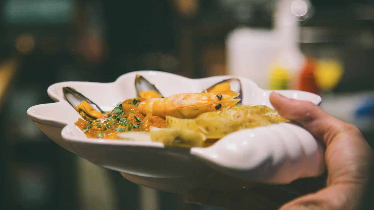Una cazuela de mariscos servida en plato hondo, sostenida por la mano de quien la lleva a la mesa